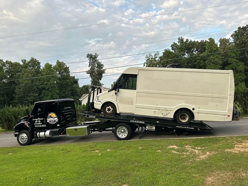 Tow truck towing a white box truck on a residential road, with trees and a cloudy sky in the background.