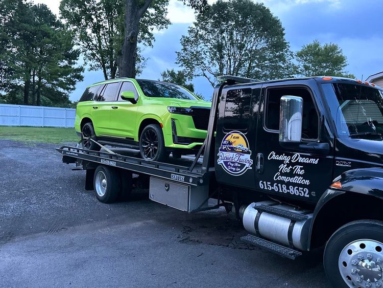 Bright green SUV on a flatbed tow truck. Black truck with logo and phone number. Outdoors.