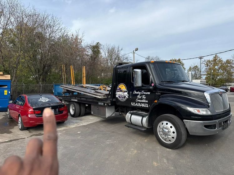 A red car being towed by a black tow truck in a parking lot. Cloudy sky.