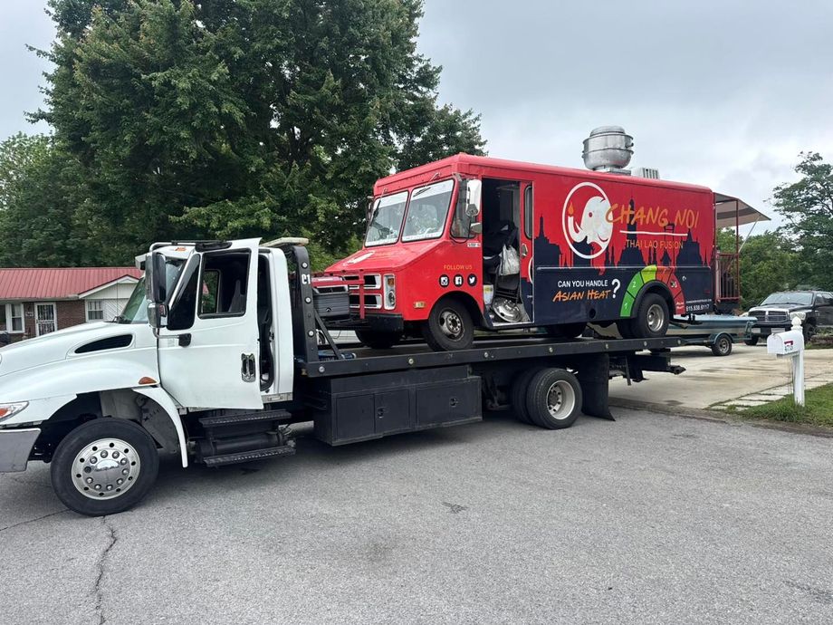 Red food truck on a flatbed tow truck in front of a house. The truck has the logo