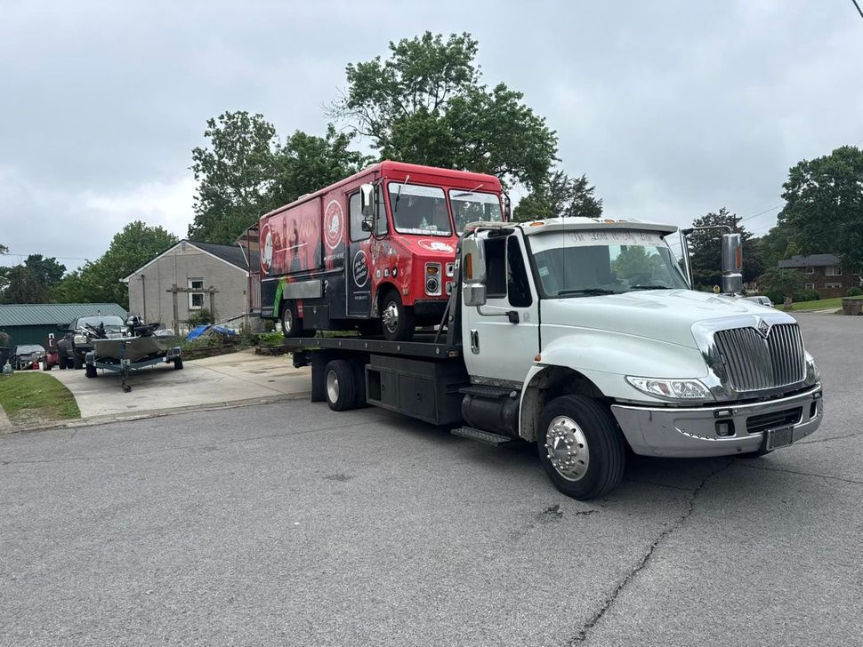 Red food truck on a white flatbed tow truck on a paved road. Cloudy day, residential area.