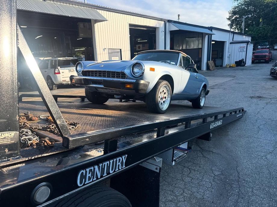 Light blue and white vintage car being towed on a flatbed tow truck in front of a repair shop.