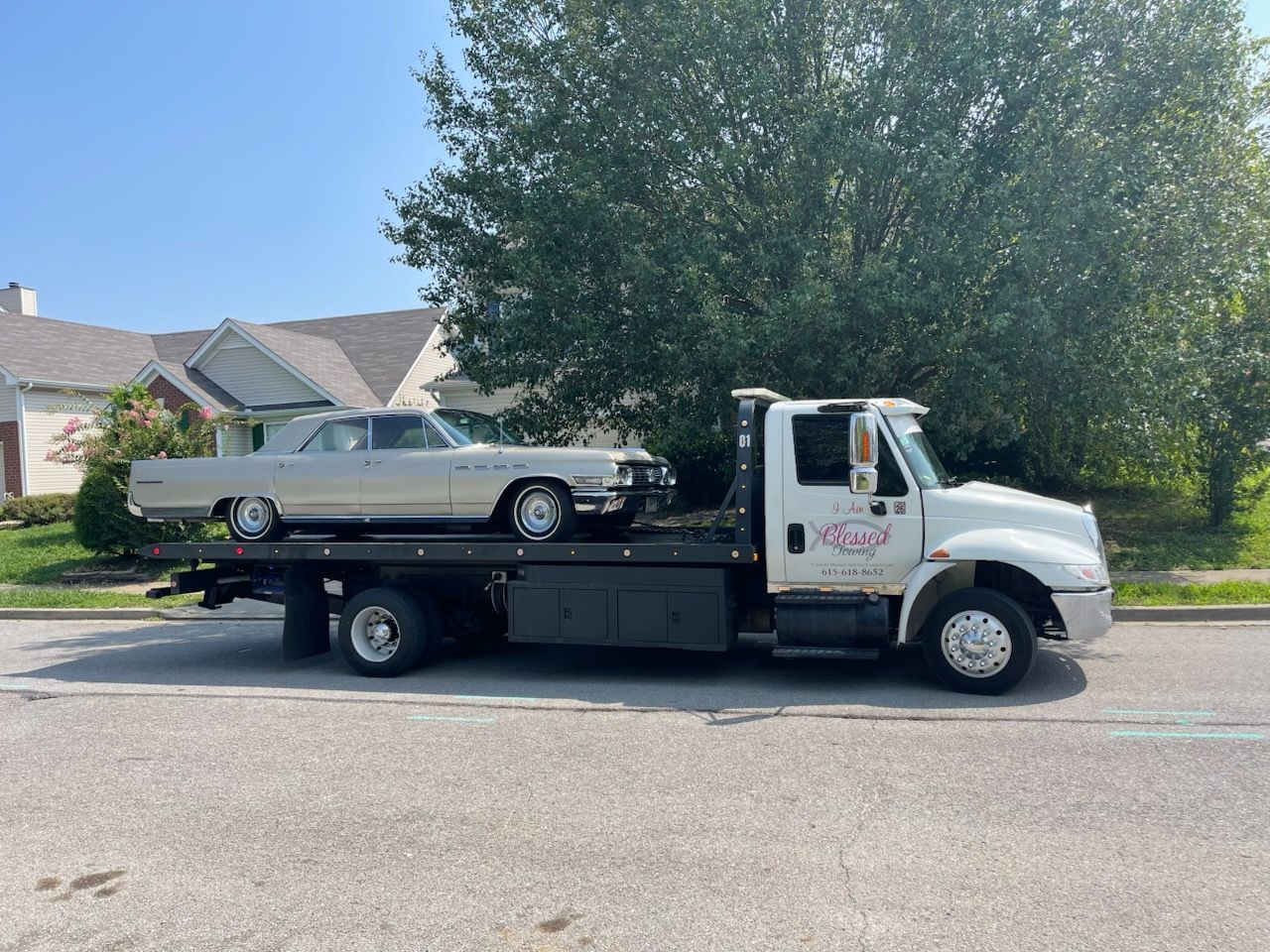 Silver classic car on a flatbed tow truck on a suburban street.