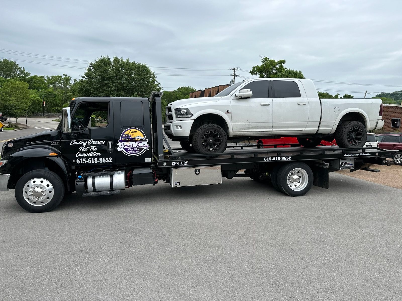 Black tow truck carrying a white pickup truck. The truck is on a flatbed, parked on pavement.