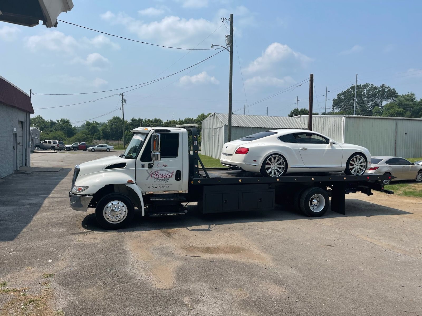 White Bentley car on a tow truck, parked near a building on a sunny day.