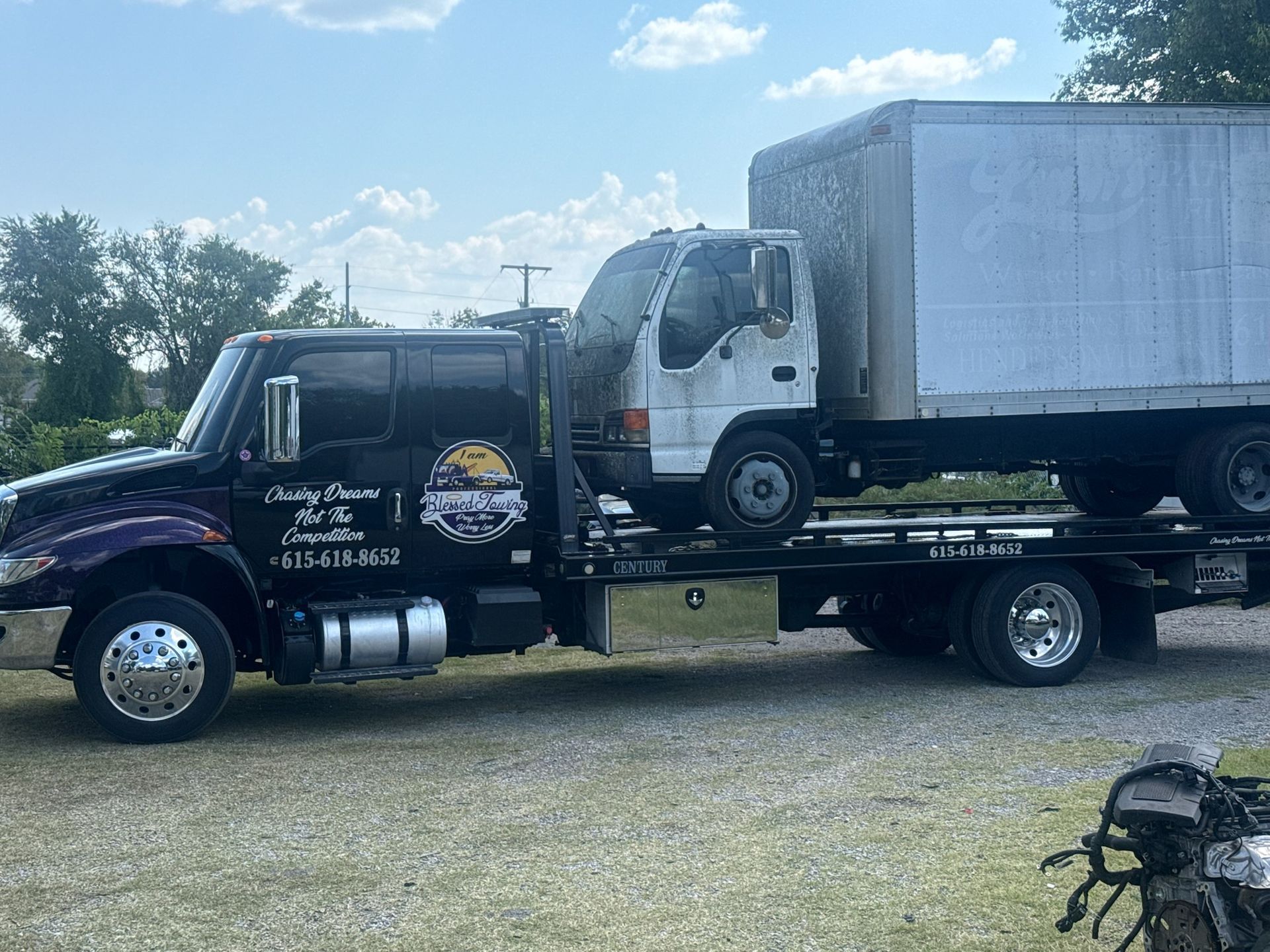 Tow truck hauling a box truck on a sunny day. The tow truck is black and purple with business name on the side.