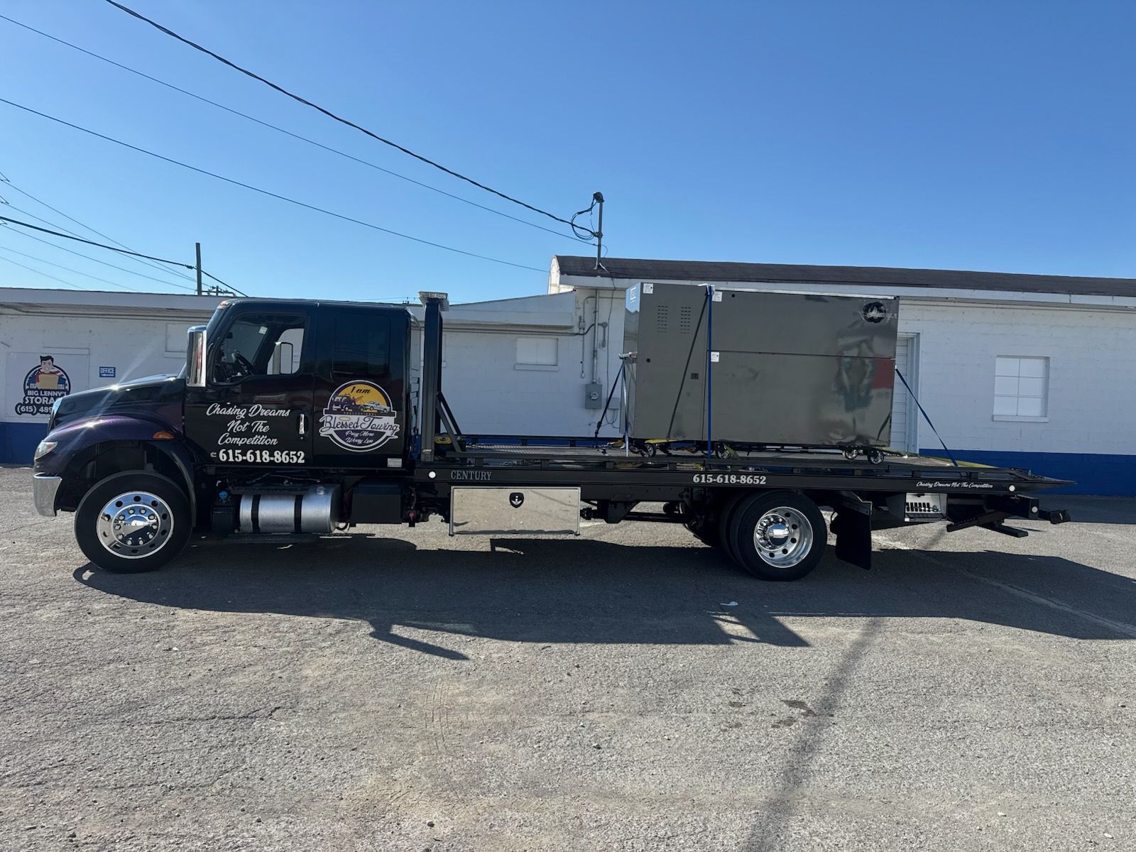 Tow truck with large box on flatbed, parked in front of a building. Sunny day.