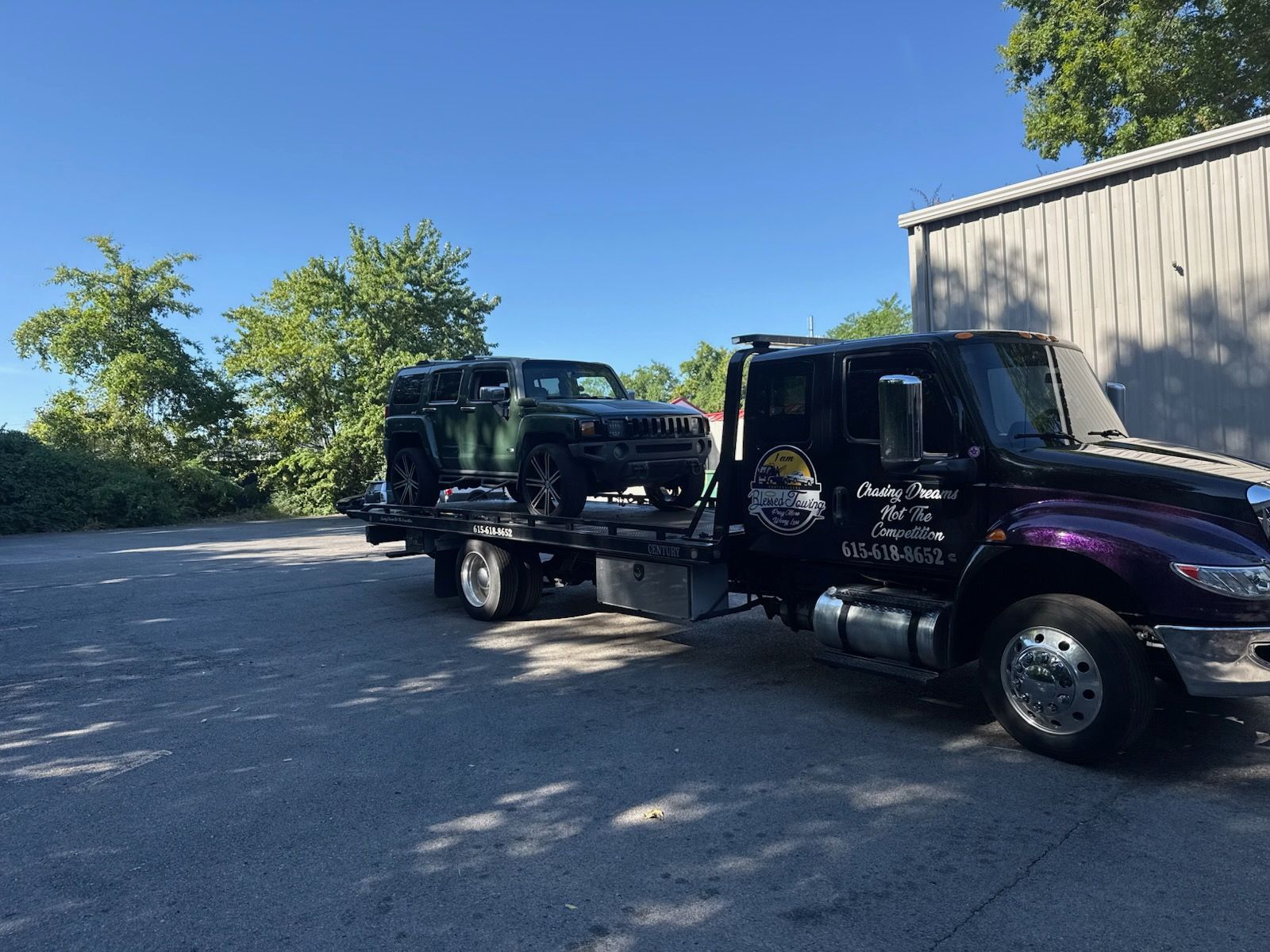 Green SUV loaded on a flatbed tow truck on a sunny day. Building and trees in the background.