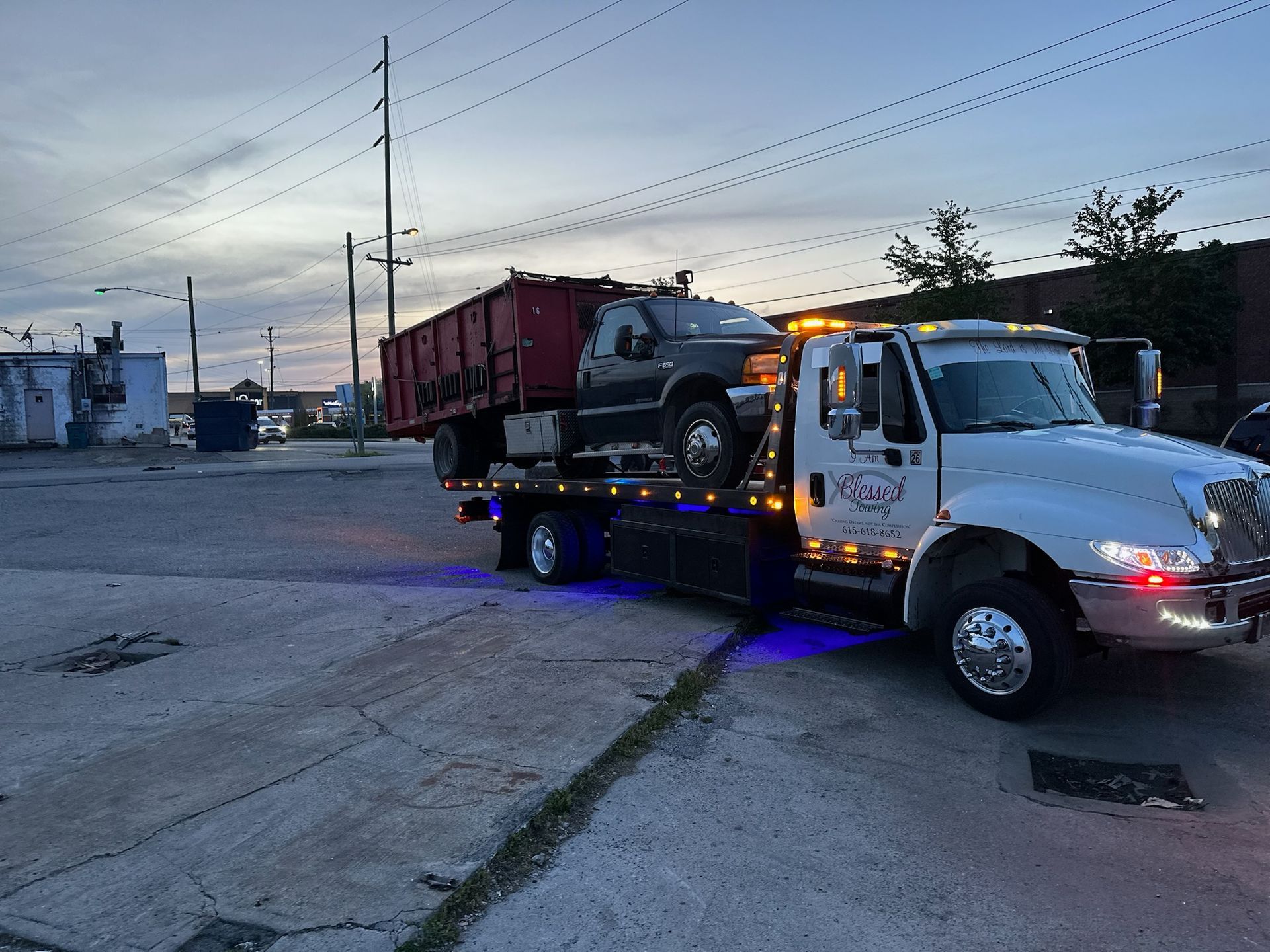 Tow truck carrying a black pickup truck and a dumpster on a paved lot at dusk.