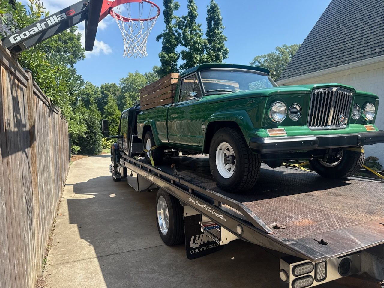Green vintage Jeep truck loaded on a flatbed tow truck next to a wooden fence.