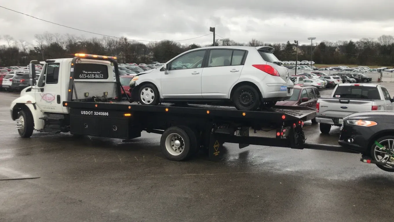 Tow truck carrying a white car on its flatbed, with another car being towed behind. Overcast day.