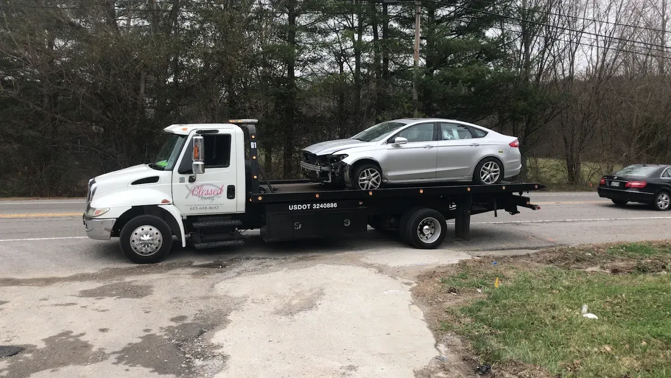 Tow truck carrying a damaged silver car on a roadside.