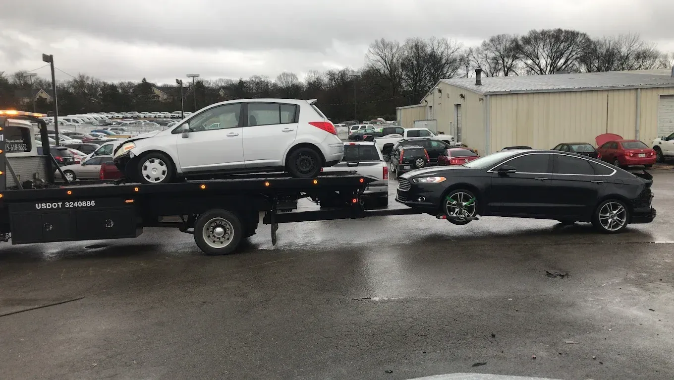 Tow truck carrying two cars in a lot. The tow truck is black and white, the cars are white and black. Overcast day.