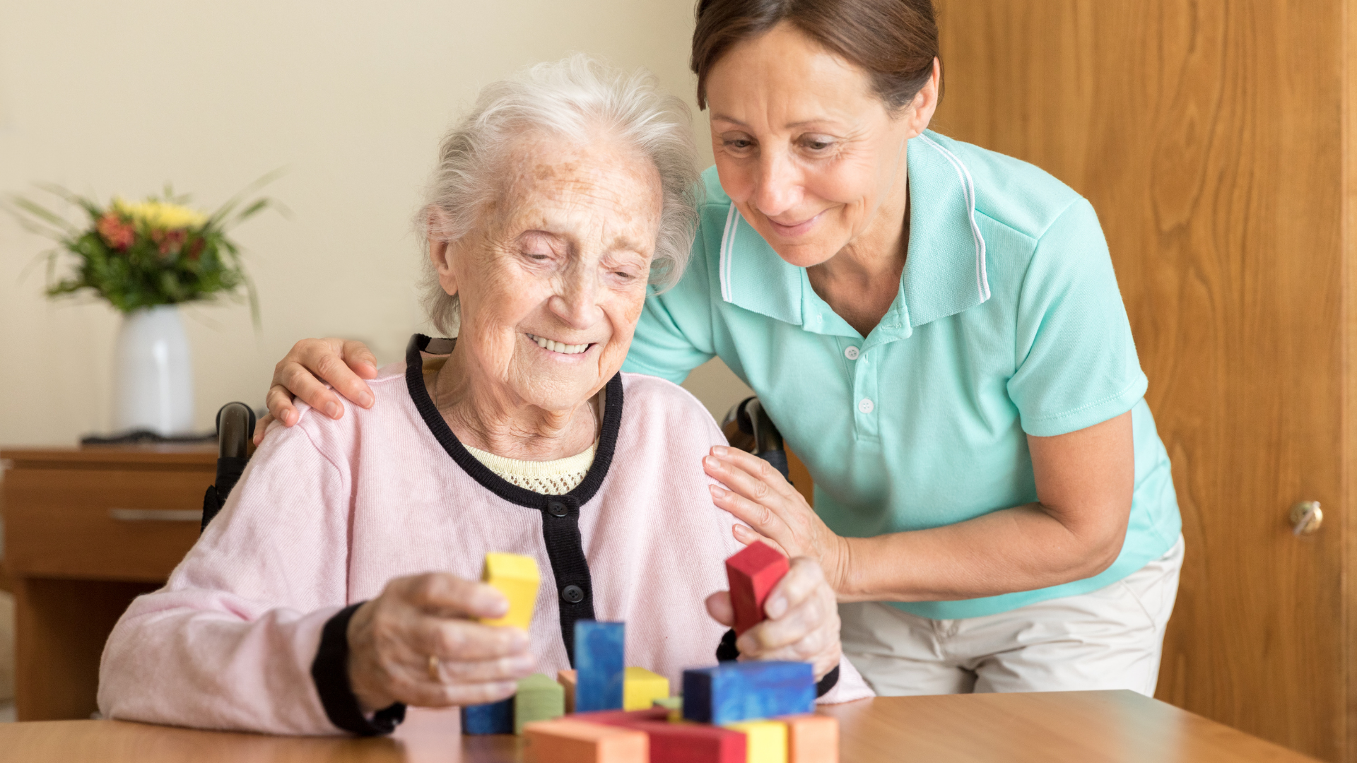 Caregiver leaning over smiling elderly woman playing with colorful blocks at a table