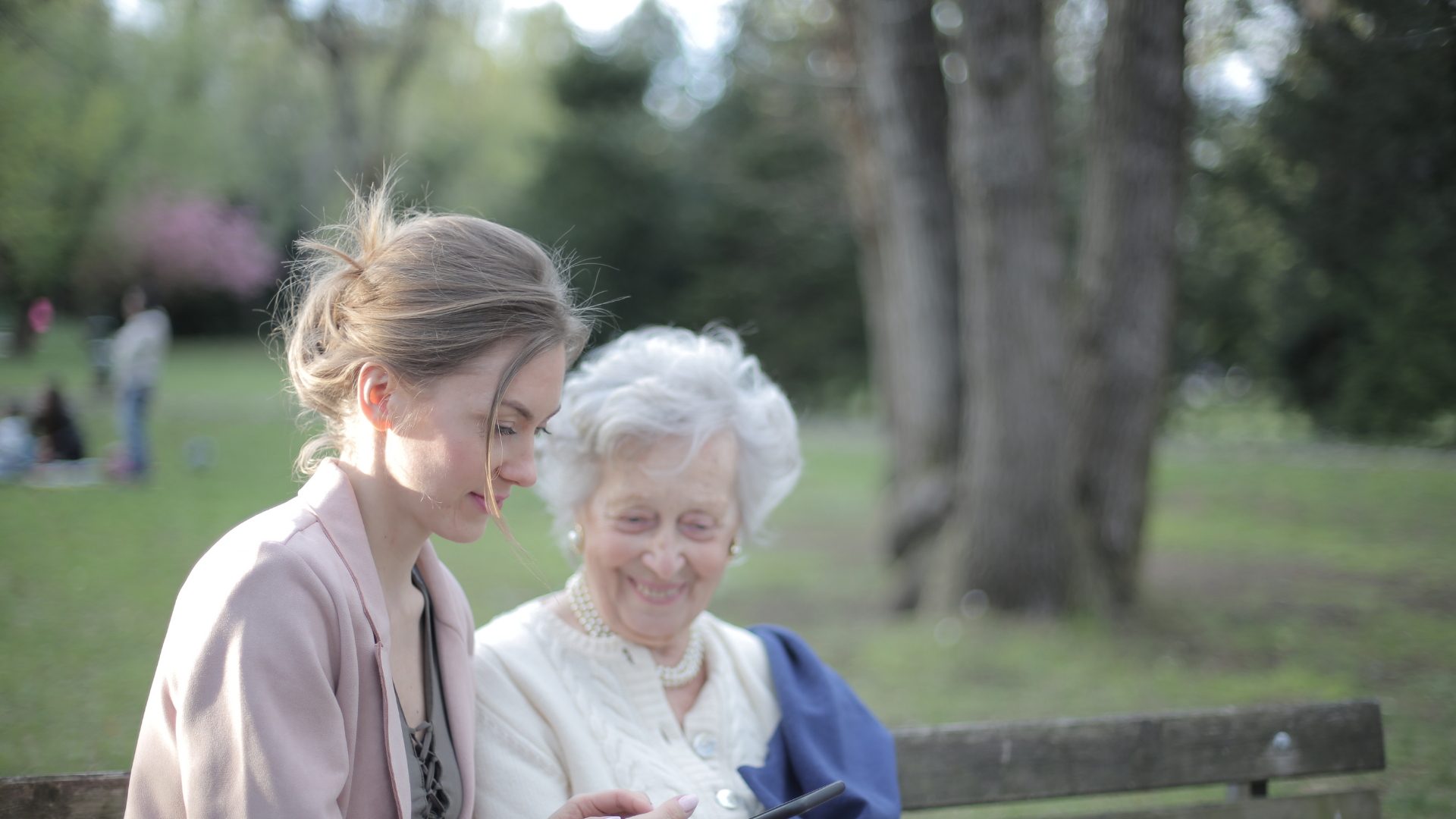 Nurse assists patient using a walker in a home. The patient wears glasses and pink shirt. The nurse smiles.