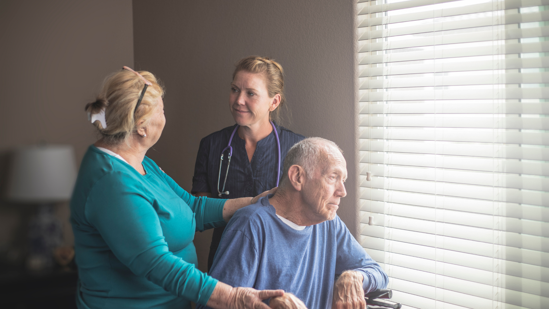 A caregiver in blue scrubs assists an older person in a white polo shirt as they stand up in a bright, modern living room.