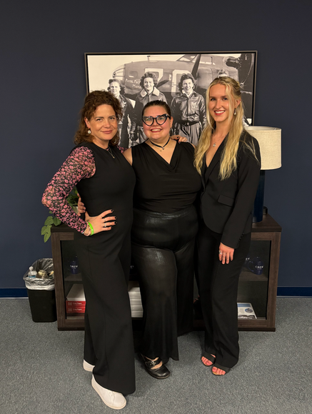 Three women are posing for a picture in front of a framed picture.