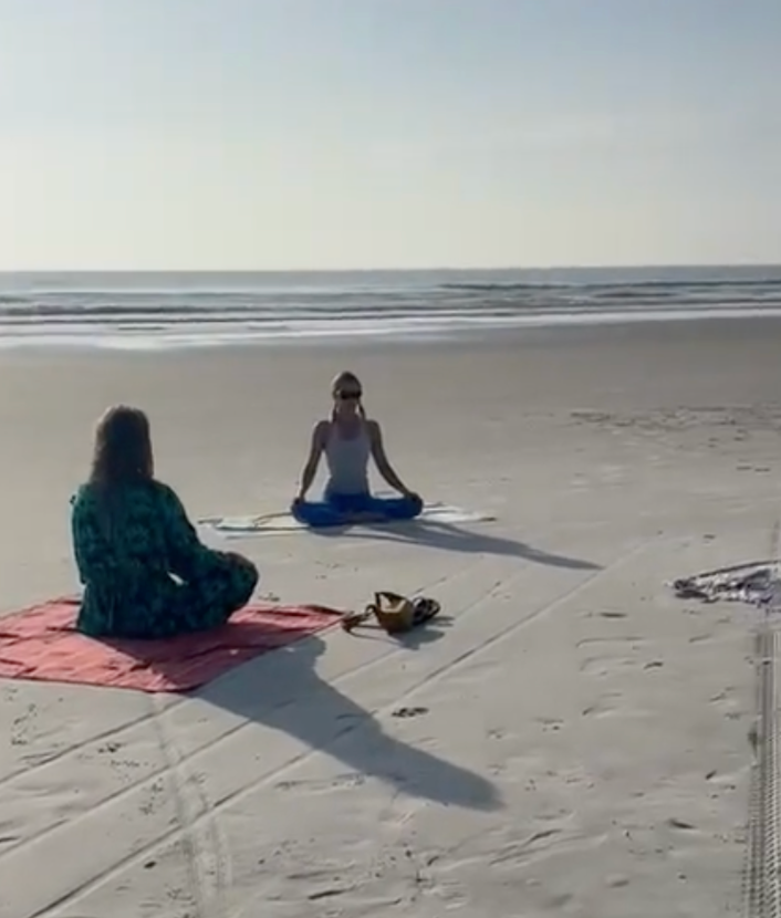 Two women are sitting on a yoga mat on the beach.