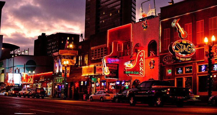 Nighttime view of neon-lit Broadway in Nashville, with bars, cars, and a glowing dusk sky.