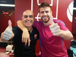 Two men, one with arm around the other, giving thumbs up; in locker room. One man has bald head and black shirt. Other in pink.