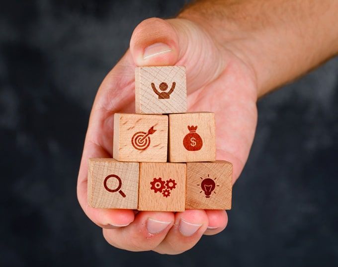 Mano sosteniendo bloques de madera apilados en forma de pirámide. Cada bloque tiene un icono diferente que representa los pasos del negocio.