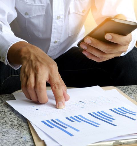Persona con camisa blanca señalando un gráfico en papel, sosteniendo un teléfono y analizando datos.