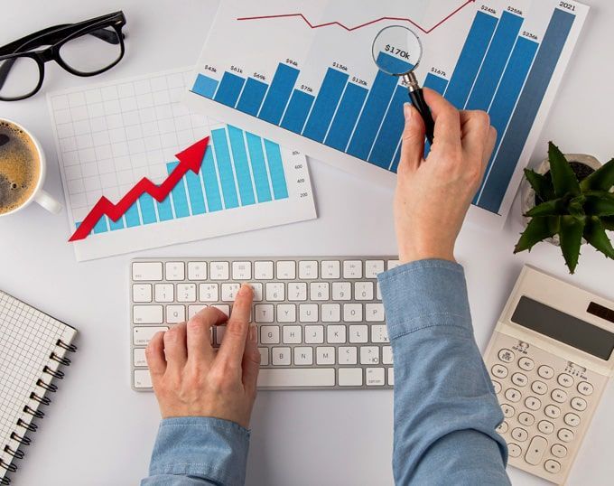 Manos utilizando un teclado, apuntando a gráficos con gráficos rojos y azules, con café, vasos y calculadora sobre un escritorio blanco.