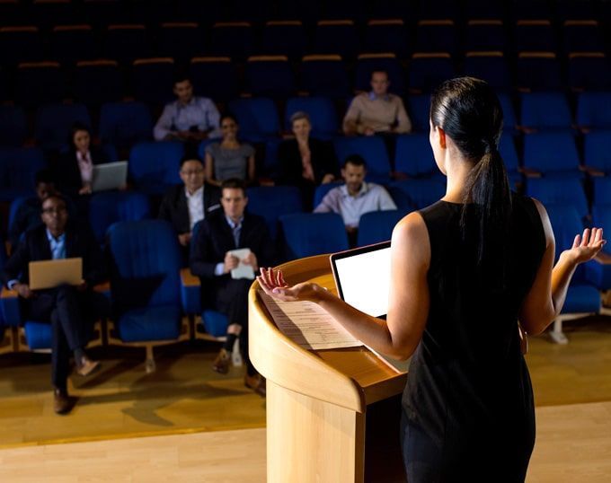Mujer presentándose en un podio ante una audiencia en un auditorio, gesticulando y mirando hacia la audiencia.