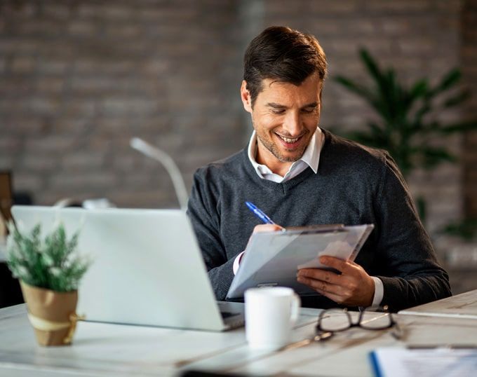 Hombre sonriendo, escribiendo en un portapapeles en un escritorio con computadora portátil, taza, vasos y una pequeña planta en la oficina.