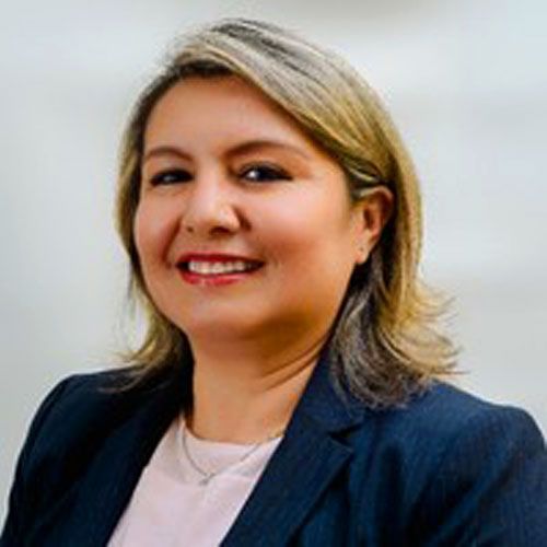 Mujer con cabello rubio rizado sonriendo, vistiendo una camiseta blanca y un collar de oro, al aire libre.