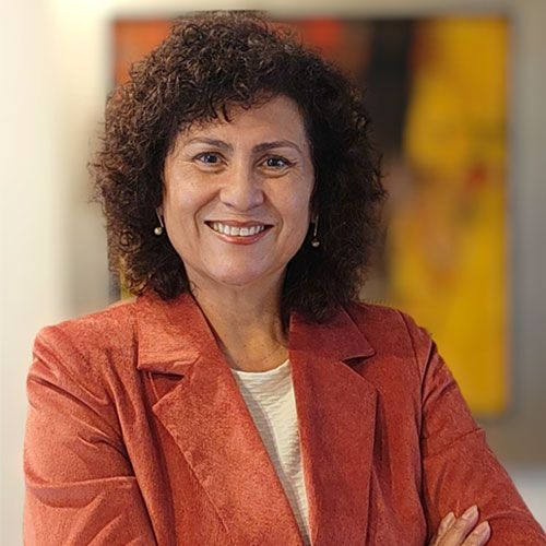 Mujer con cabello rubio hasta los hombros, con top azul, apoyando la barbilla en la mano, sonriendo.