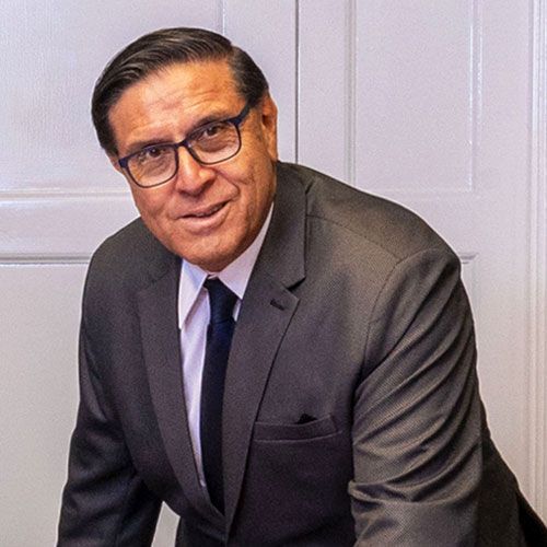 Hombre de traje y gafas, sonriente, apoyado en una pared blanca.