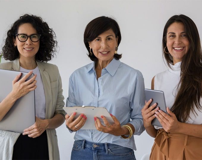 Tres mujeres sonrientes, de distintas edades, con portátiles, portapapeles y tabletas. Fondo blanco.