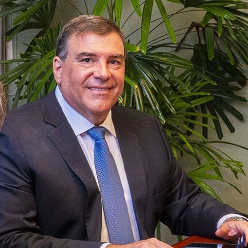 Hombre con traje sonriendo, en un espacio interior, con plantas frondosas al fondo.