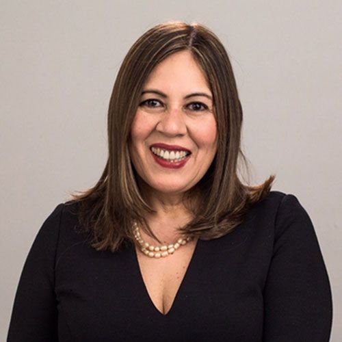 Una mujer con cabello rubio hasta los hombros sonríe, viste un blazer azul, una camisa rosa y un collar.