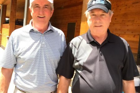 Two men are standing next to each other in front of a wooden building.