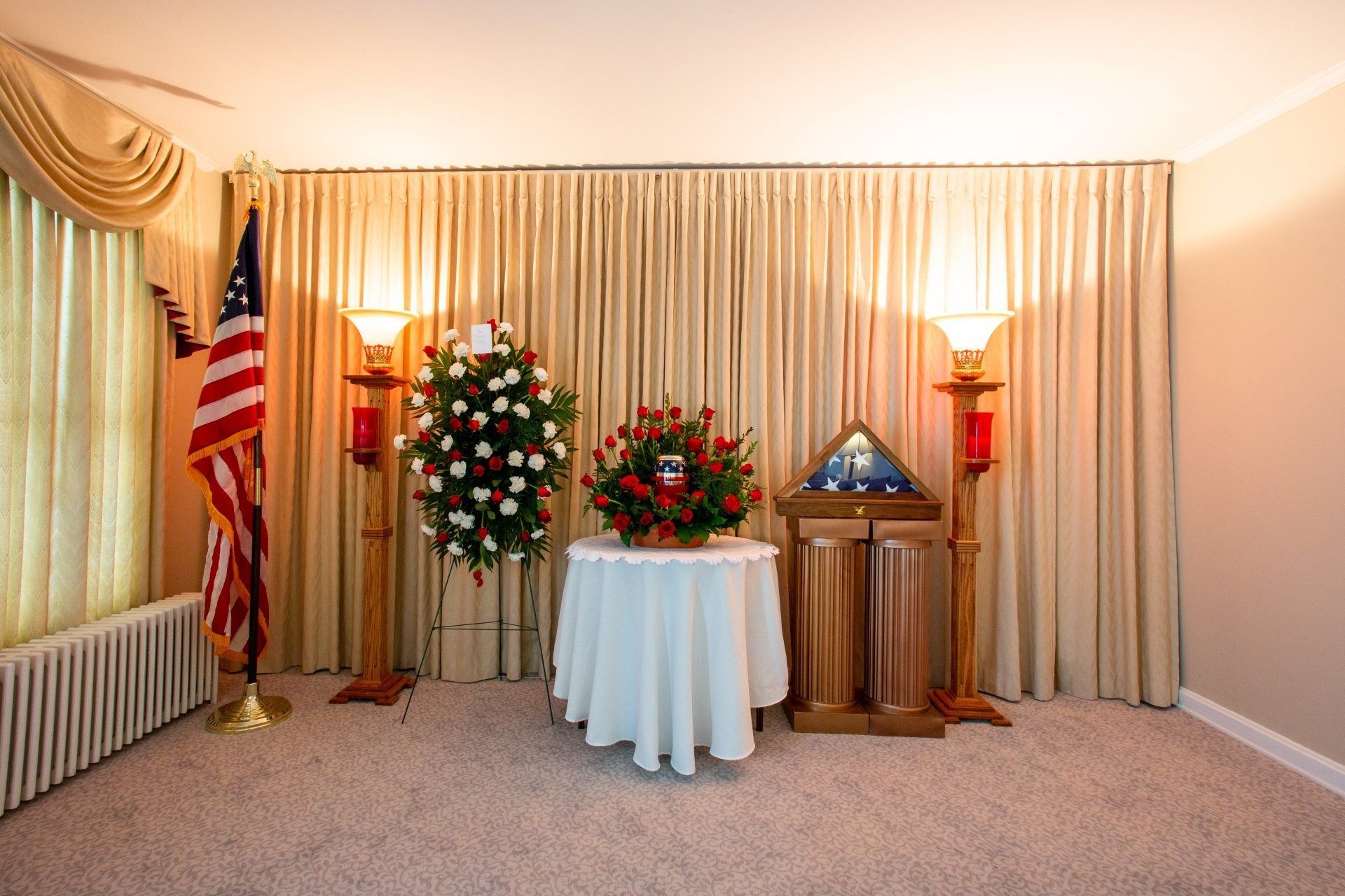 Memorial display with American flag, floral arrangements, and draped curtains in a carpeted room