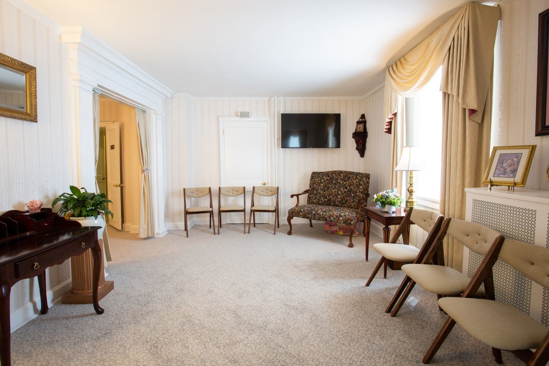 Bright living room with beige carpet, cream drapes, chairs, a table, and a wall-mounted TV.