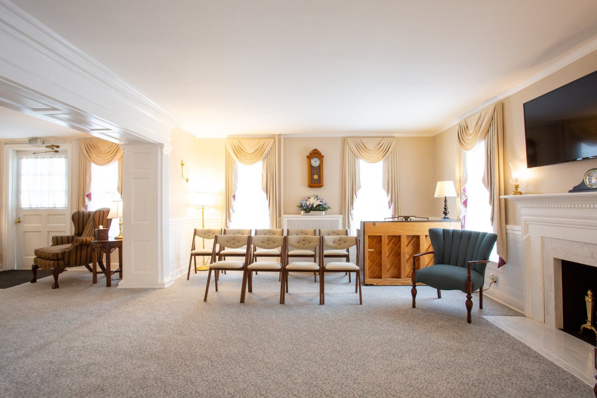 Bright living room with carpet, dining table, fireplace, wall TV, and sunlit windows with drapes