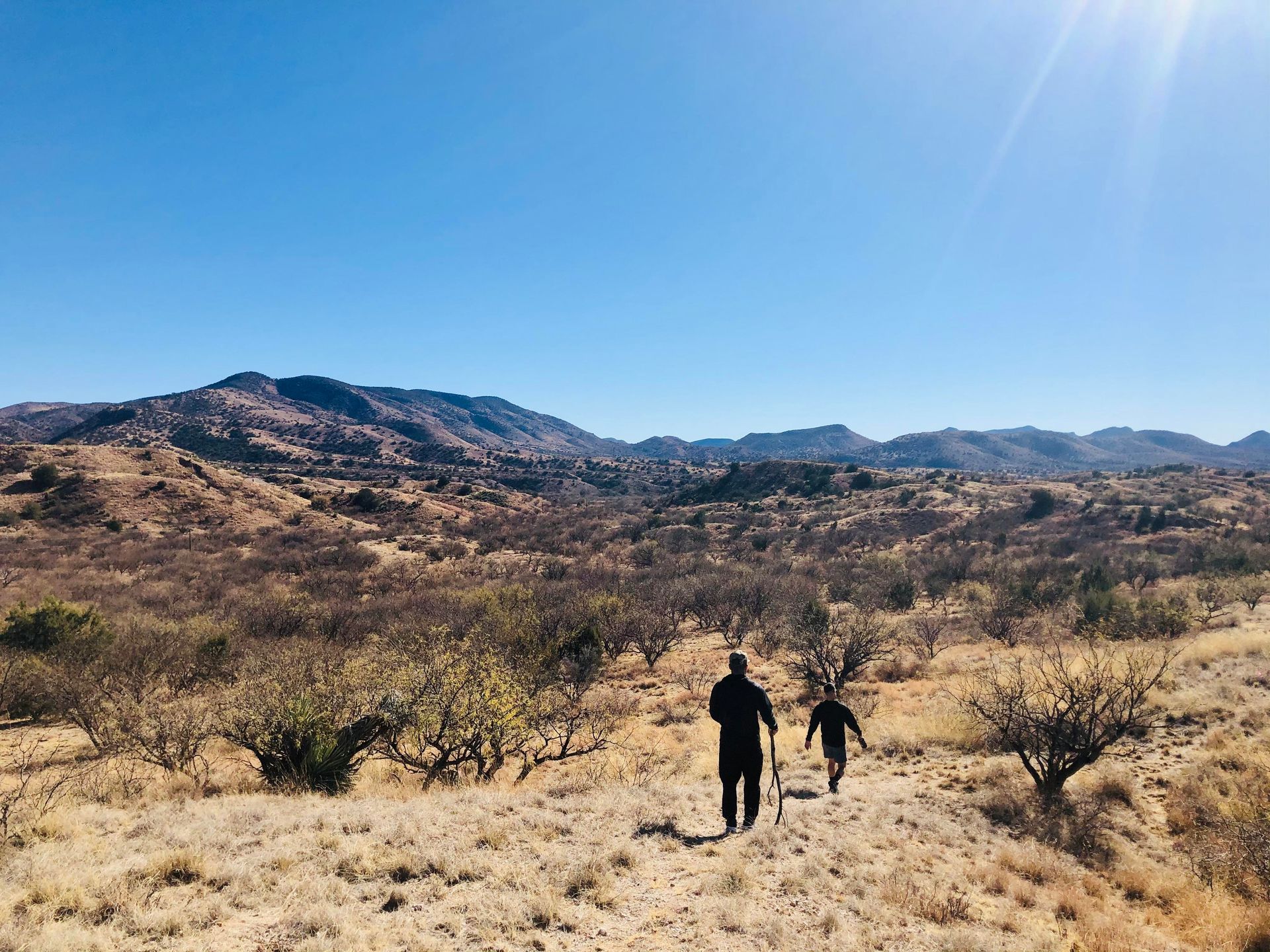 Two people are walking in the desert with mountains in the background.