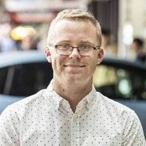A man wearing glasses and a white shirt is smiling in front of a car.