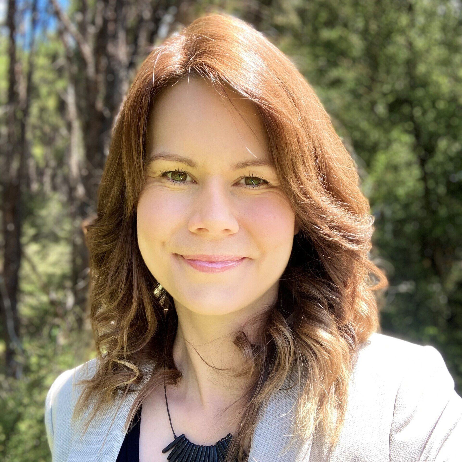 A woman with red hair is smiling for the camera in front of a forest.