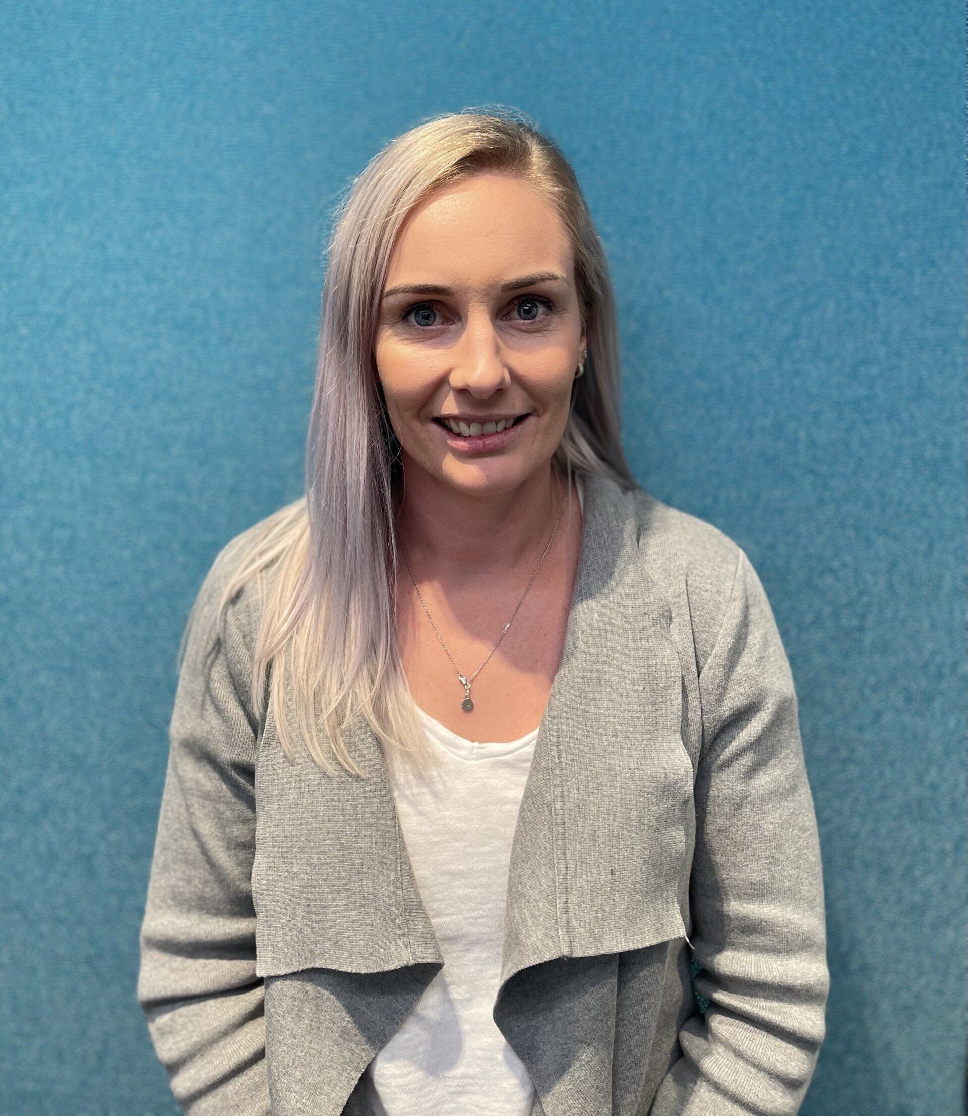 A woman wearing a grey jacket and a white shirt is standing in front of a blue wall.