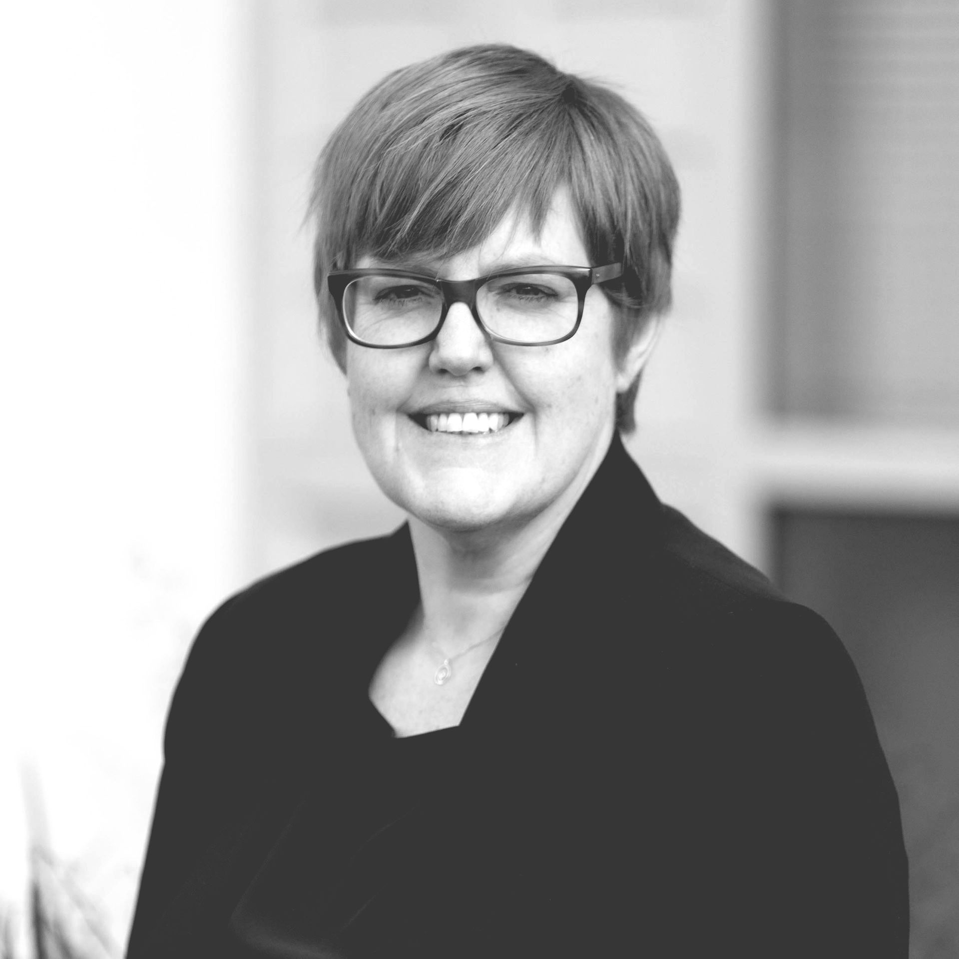 A woman wearing glasses and a black jacket is smiling in a black and white photo.