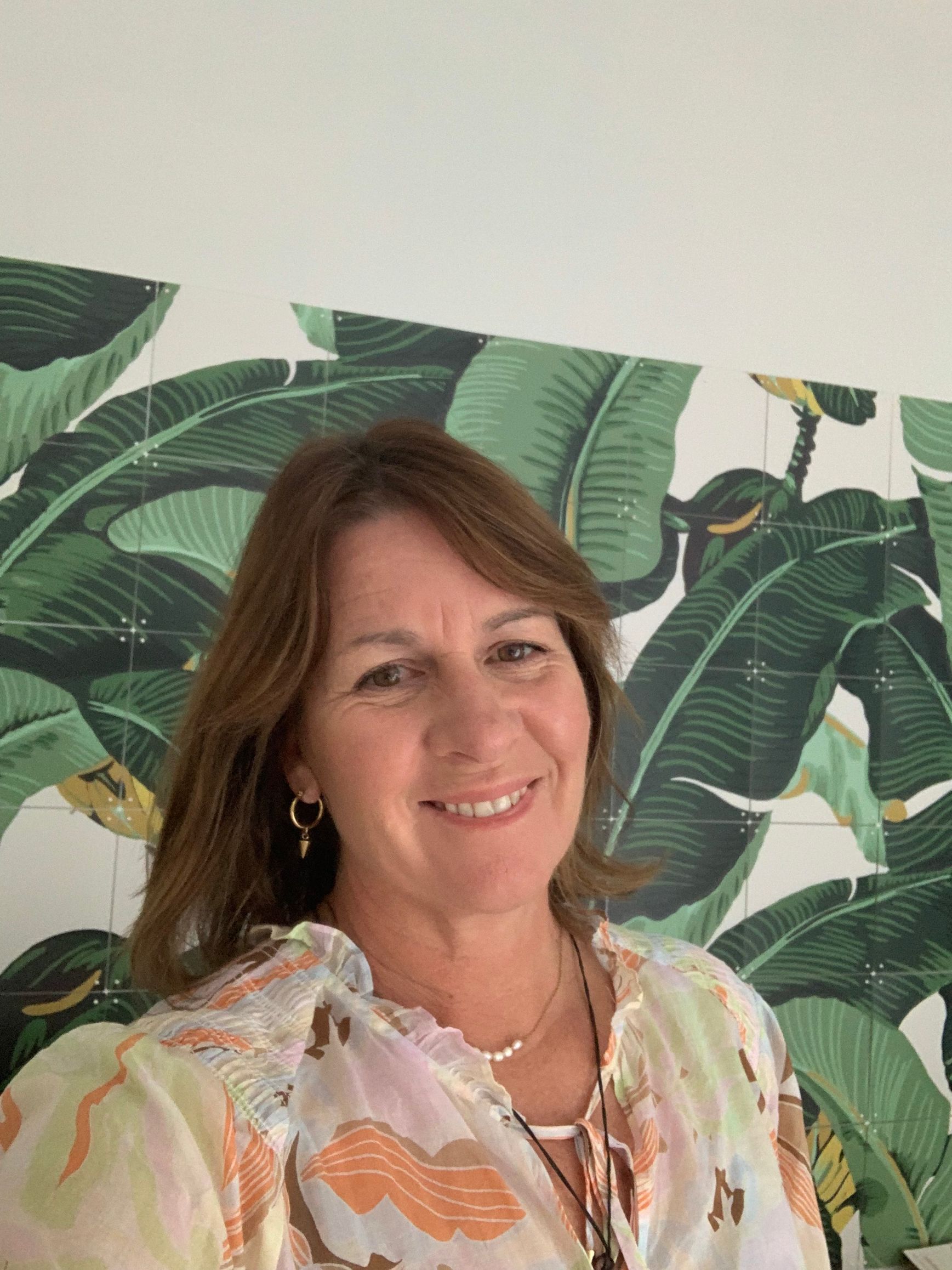 A woman is smiling in front of a wall with banana leaves on it.