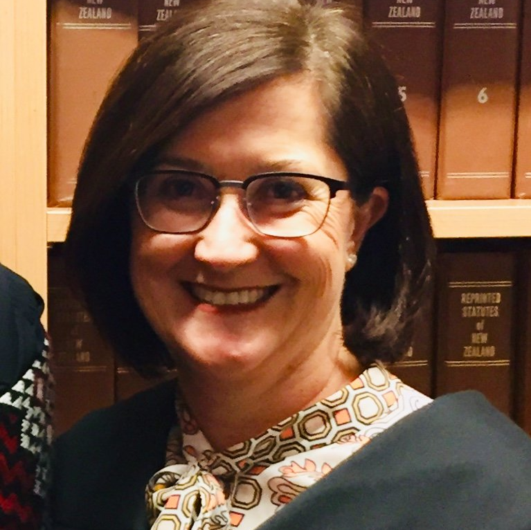 A woman wearing glasses and a scarf is smiling in front of a bookshelf with new zealand books on it