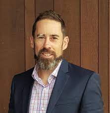 A man with a beard and a suit is standing in front of a wooden wall.