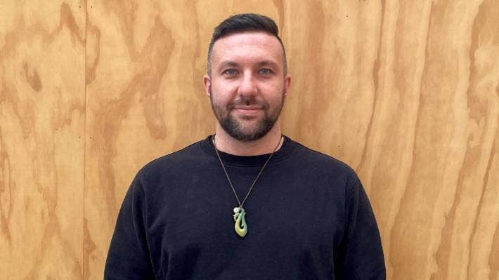 A man with a beard and a necklace is standing in front of a wooden wall.