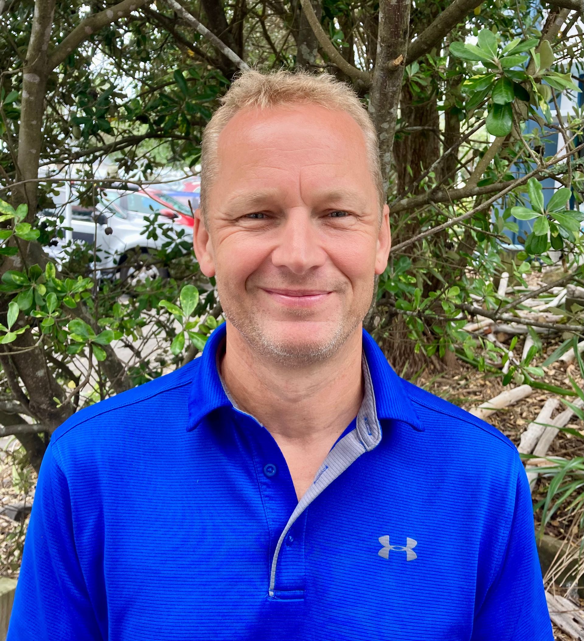A man wearing a blue under armour shirt is standing in front of a tree.