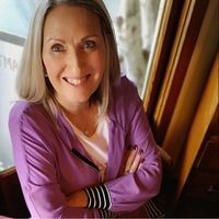A woman in a purple jacket is sitting in front of a window with her arms crossed and smiling.